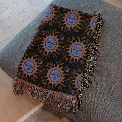 Luxurious woven cotton throw blanket featuring mystical blue sun faces with copper rays on black background, bordered by elegant fringe. Perfect celestial wall hanging, witchy bedroom decor, or dark academia throw for couch or bed.
